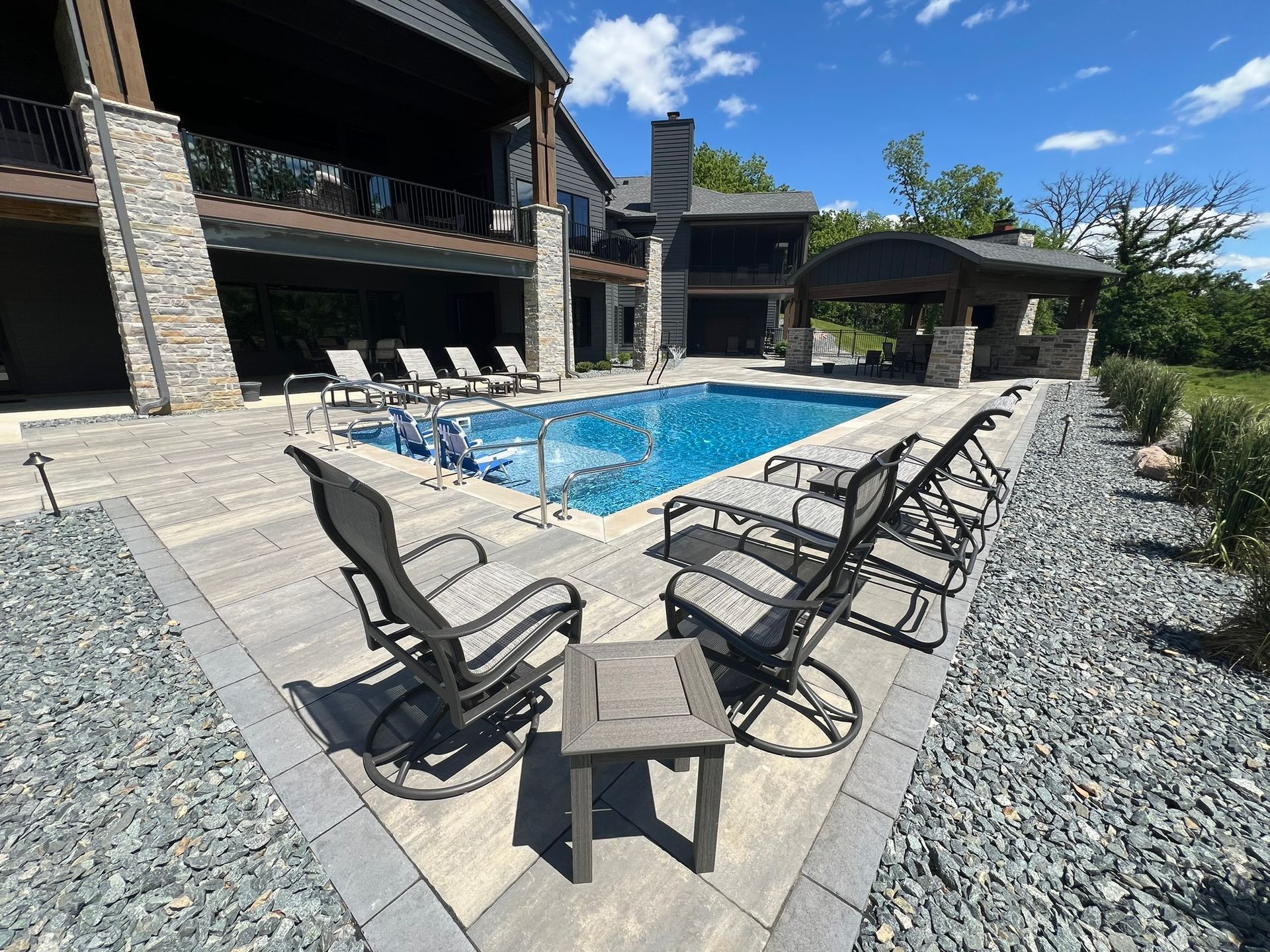 A backyard pool area with stone patio, lounge chairs, and a covered patio structure under a bright, clear blue sky.