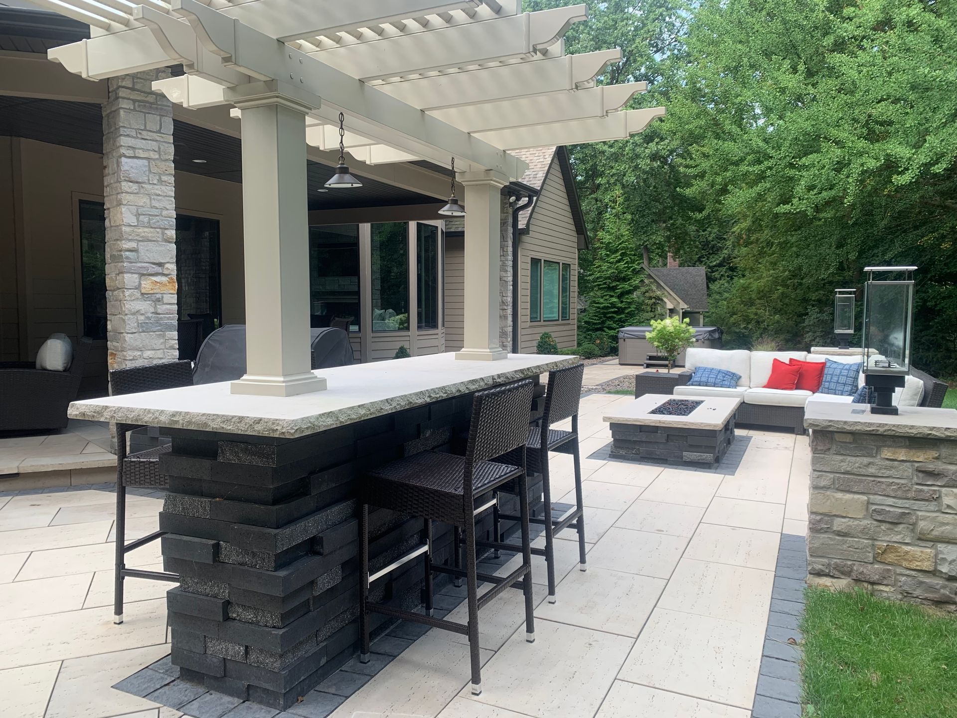 An outdoor patio features a stone bar with tall stools under a pergola, a stone fire pit, and a sofa seating area.