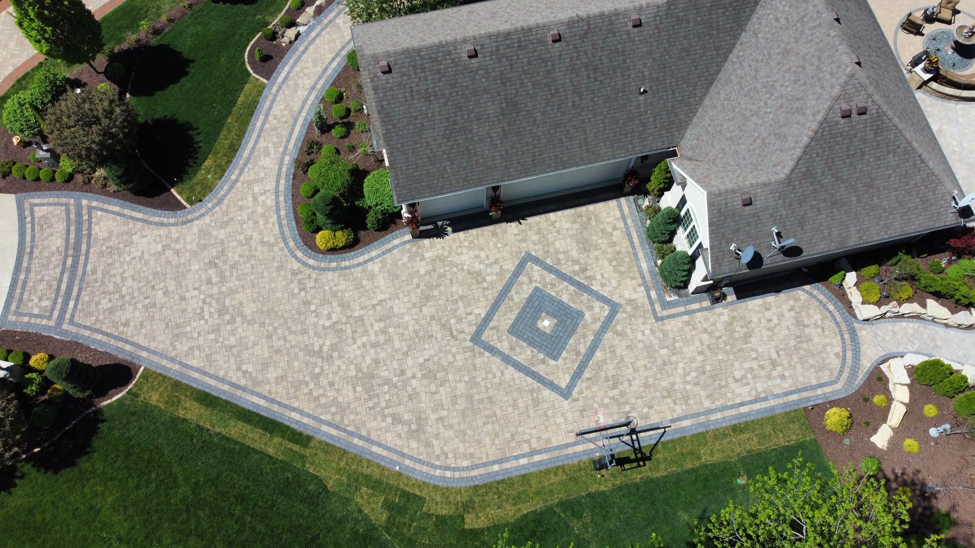 Aerial view of a paved residential driveway with a diamond-shaped accent pattern, bordered by green landscaping and a home.