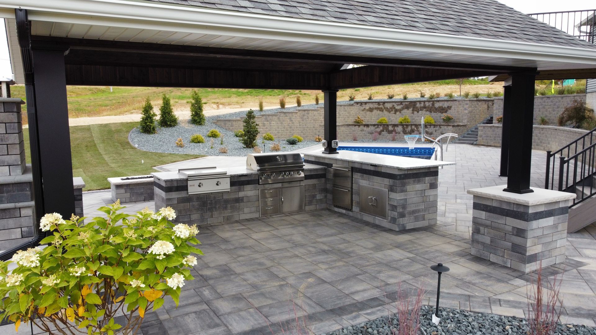 A gray stone outdoor kitchen under a roofed pavilion, featuring a grill, counter space, and a nearby swimming pool.
