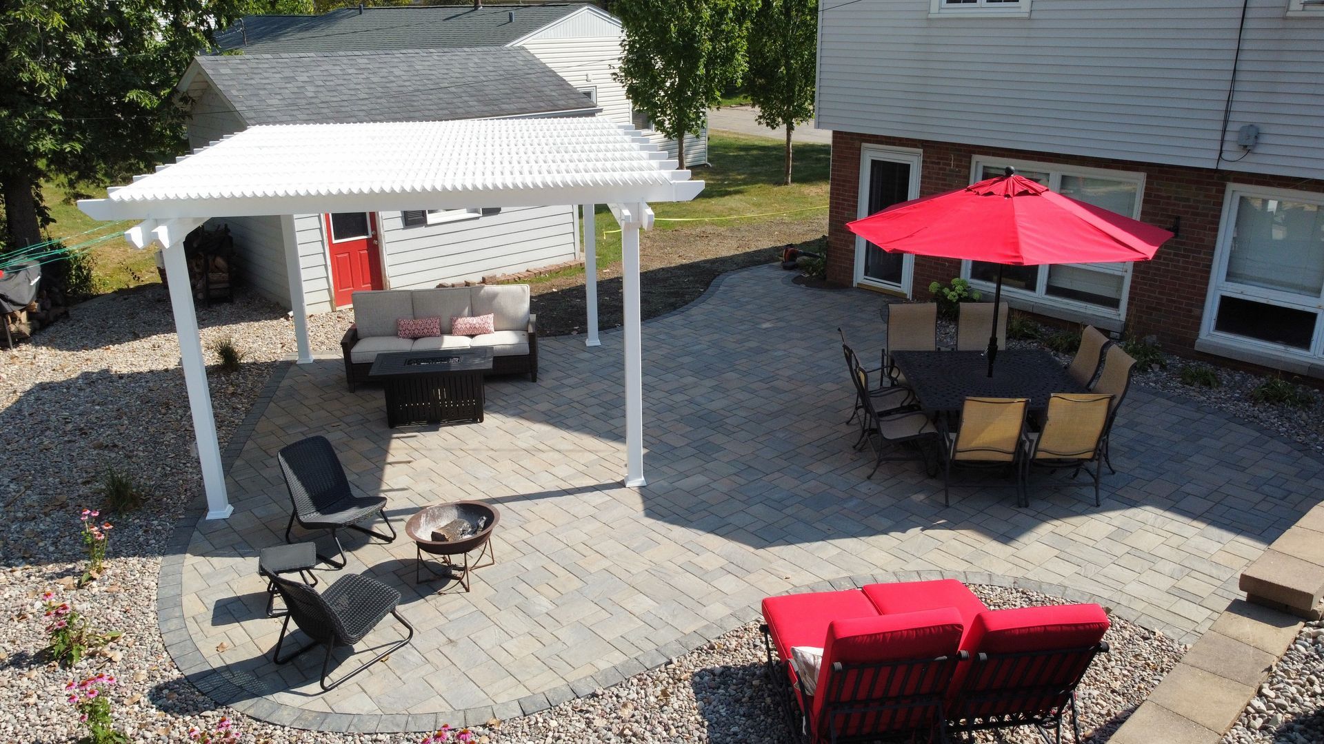 A stone patio features a white pergola over a seating area, a dining table with a red umbrella, and red lounge chairs.