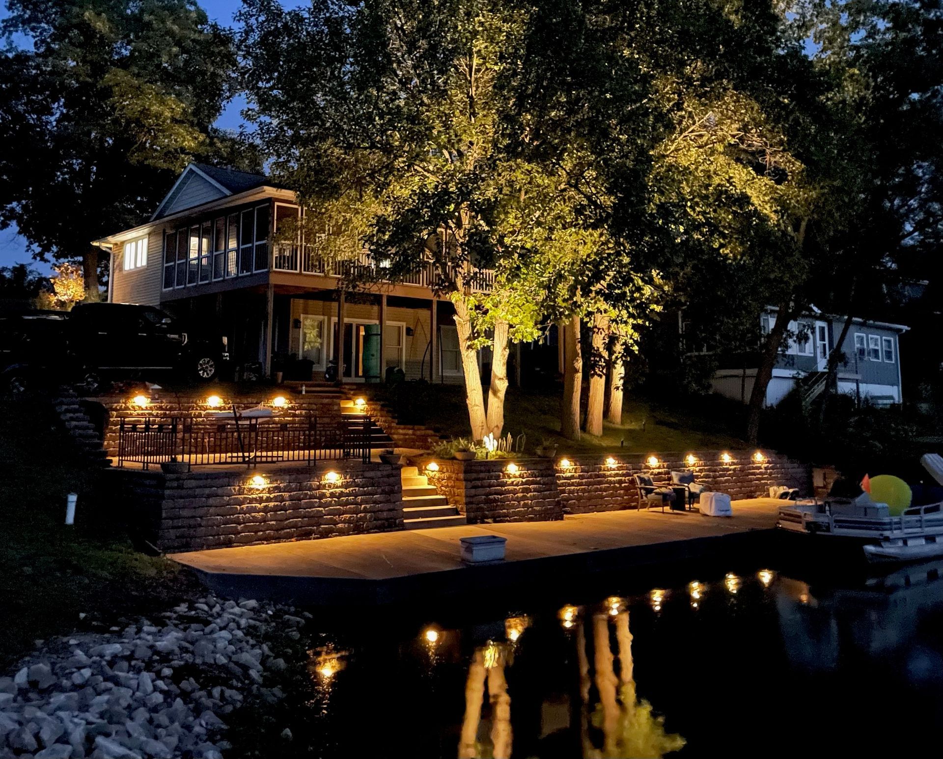 A multi-level home and stone retaining wall at night, illuminated by warm landscape lighting overlooking a lake.