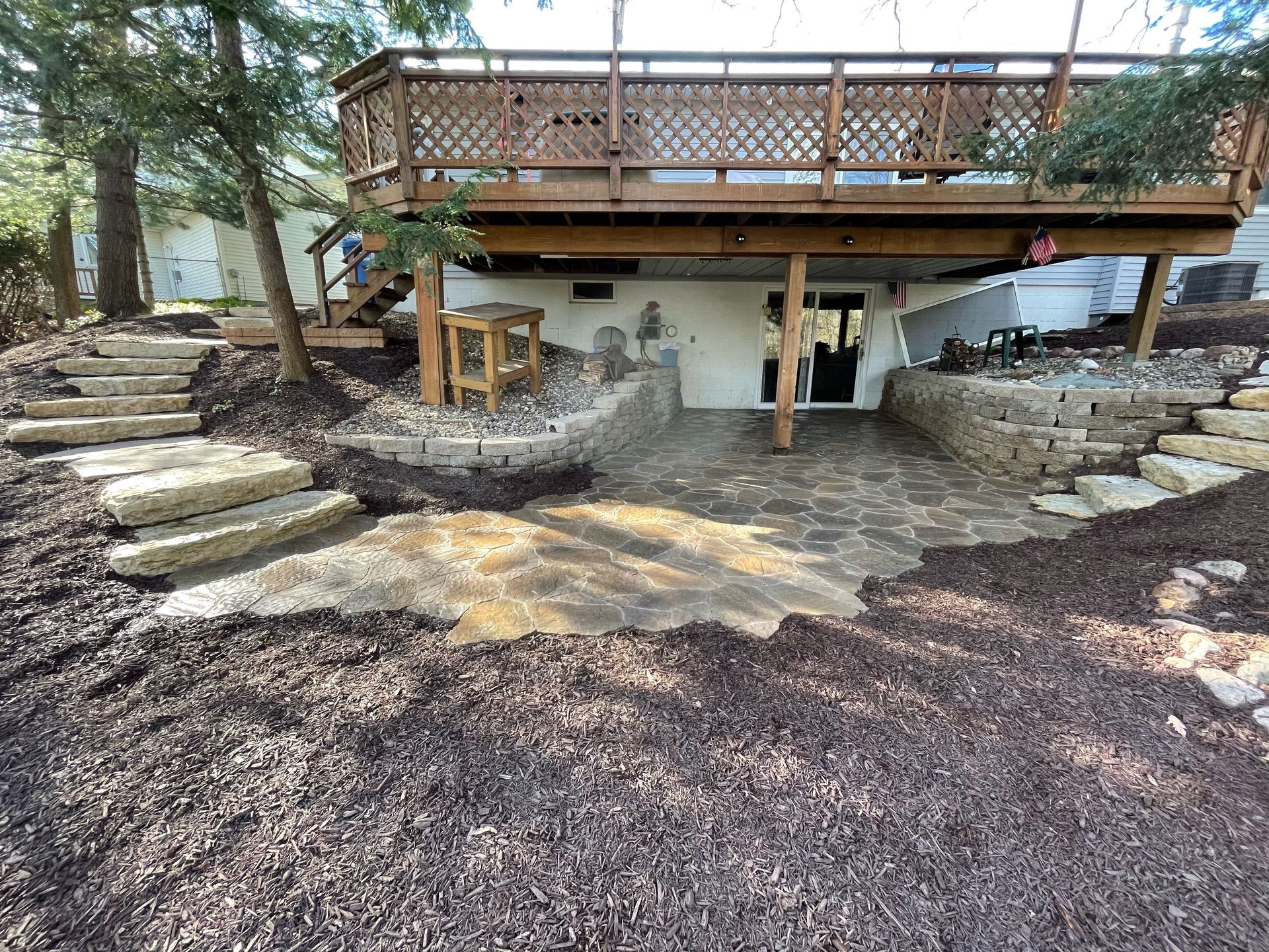 A stone patio under a wooden deck, surrounded by mulch, with stone steps leading up both sides.