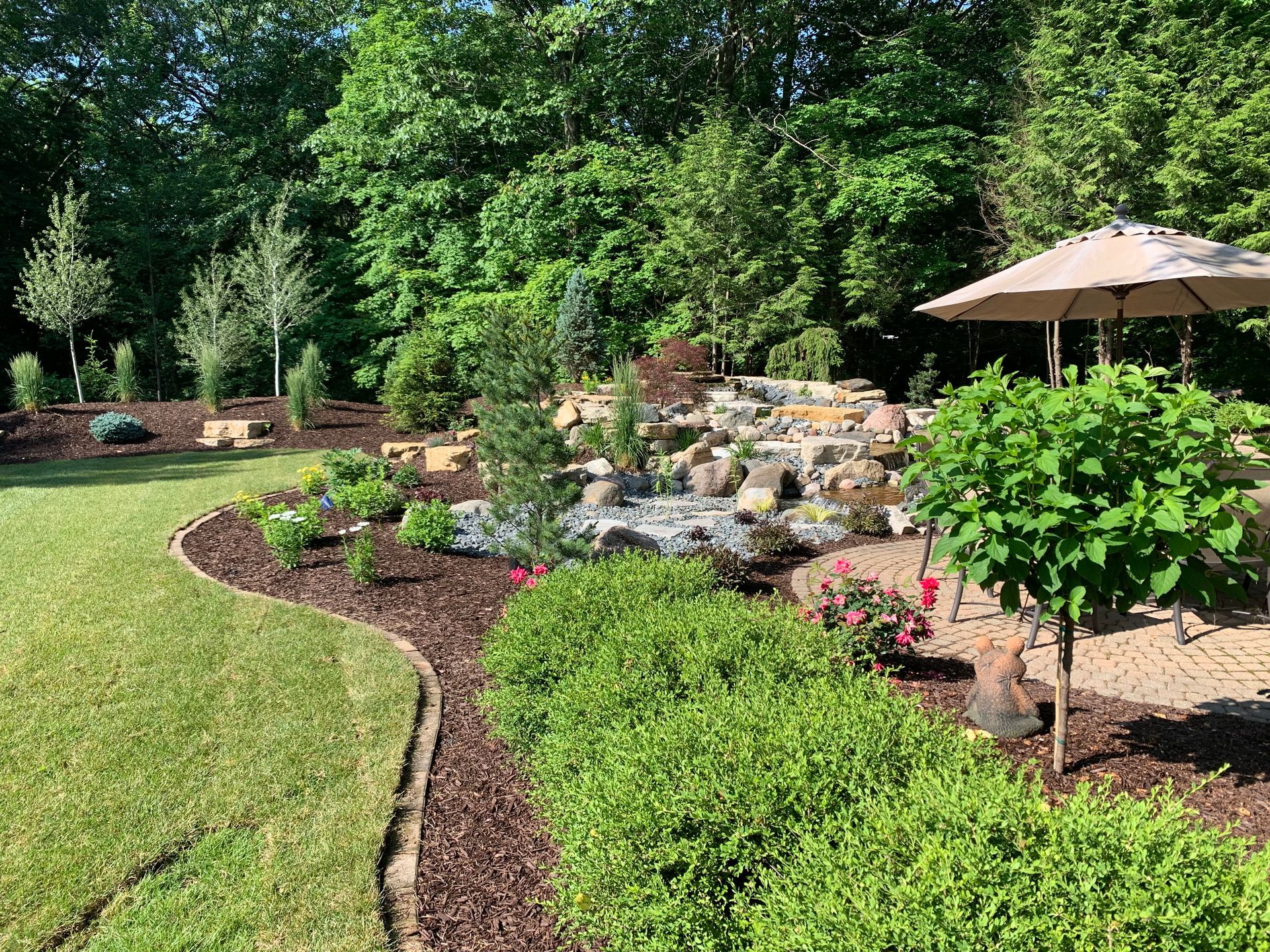 A landscaped backyard featuring a stone water feature, lush green shrubs, mulch beds, and a patio with a tan umbrella.