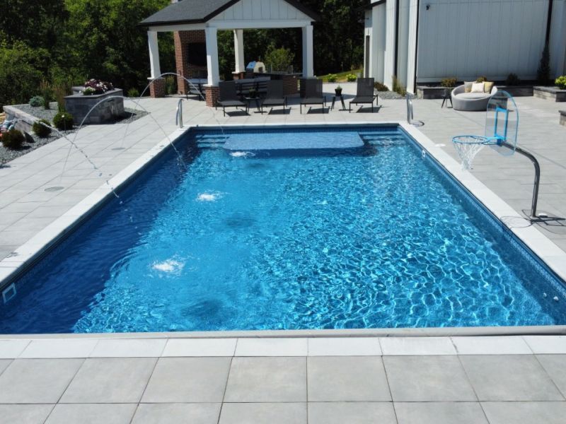 A rectangular swimming pool with blue water, water jets, a basketball hoop, and a patio area with seating.