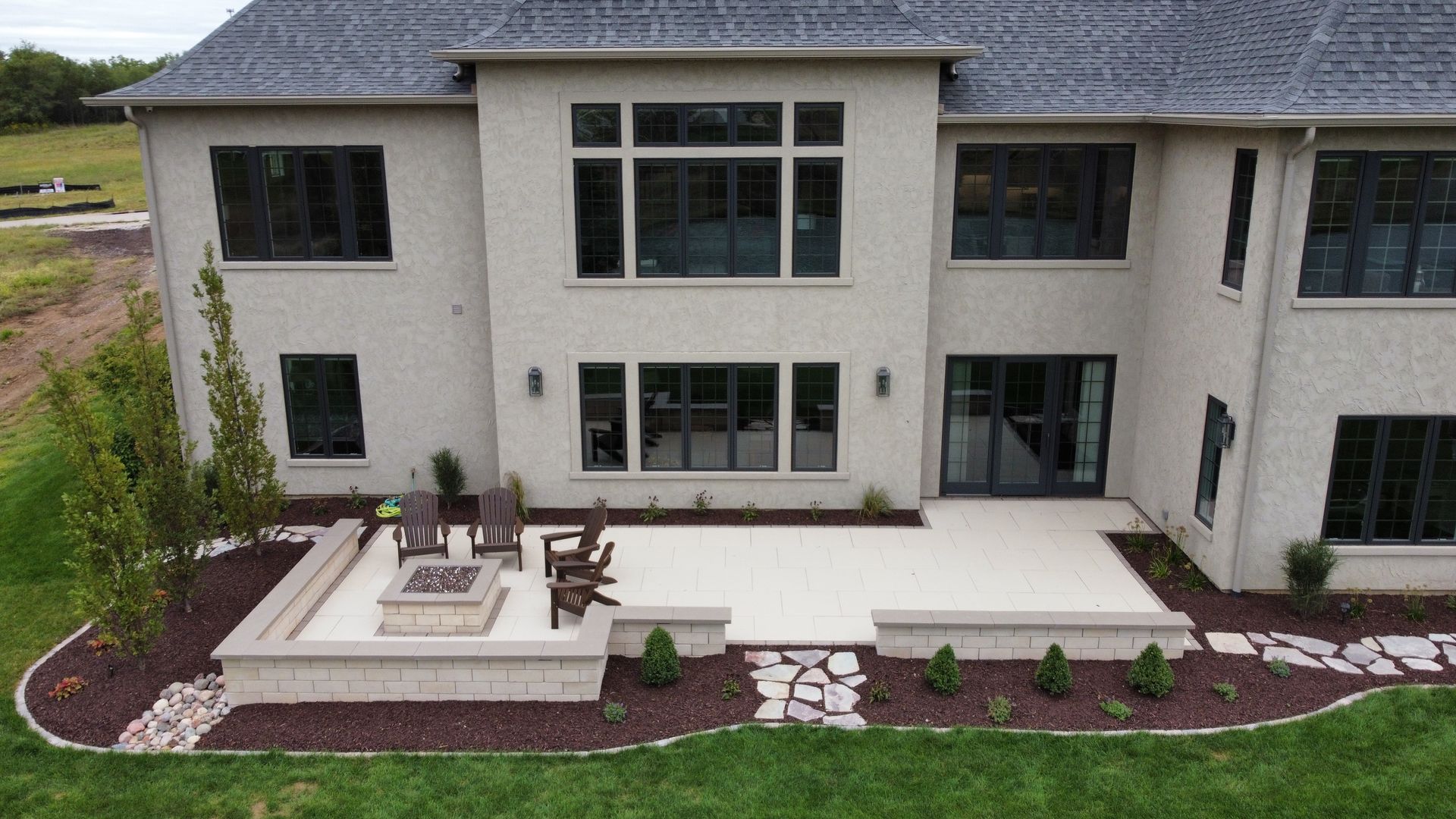 A beige stucco house features a stone patio with a fire pit, outdoor chairs, and a landscaped garden bed with mulch.