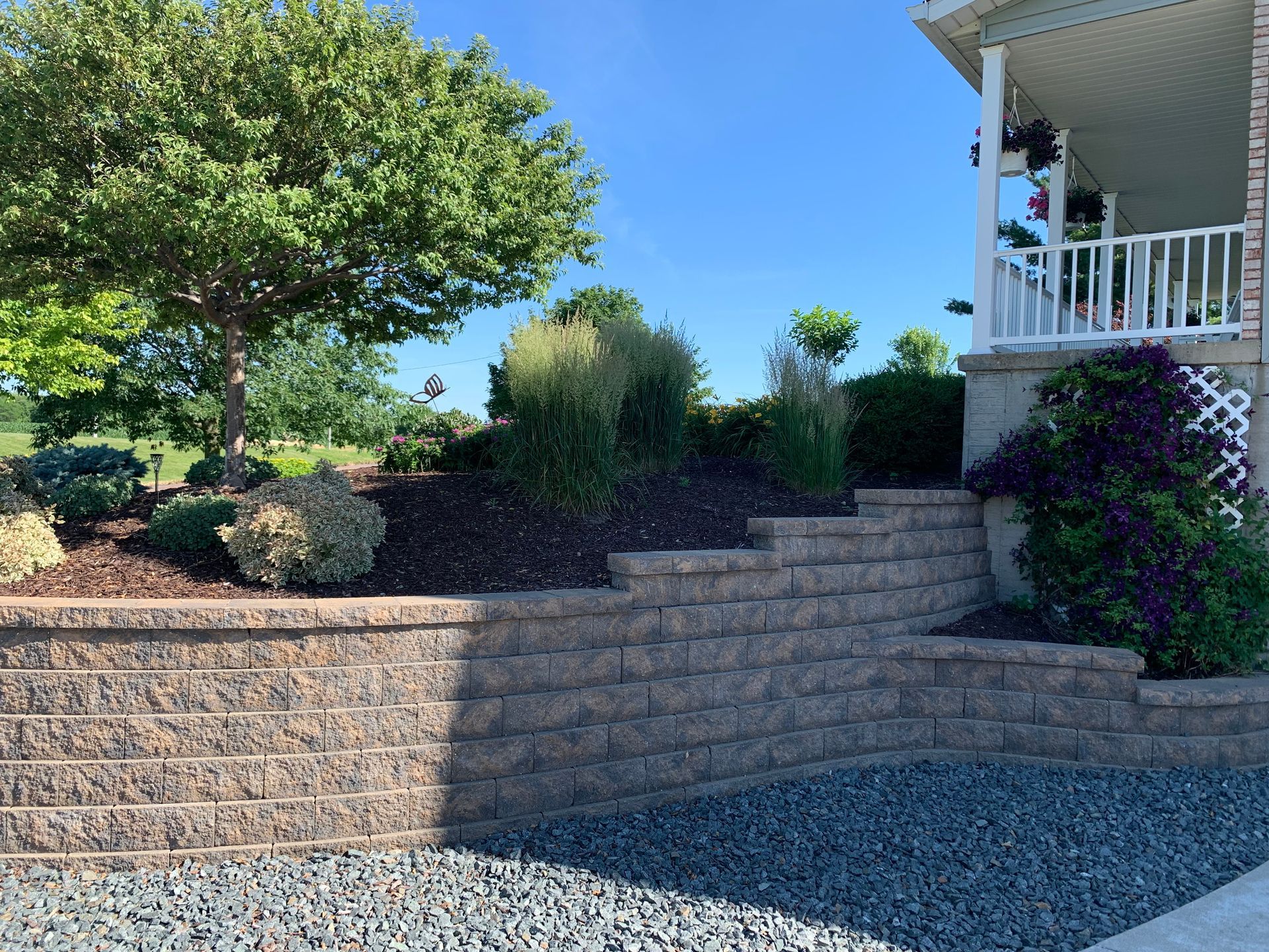 A tiered stone retaining wall landscaped with shrubs and ornamental grasses beside a house porch under a clear blue sky.