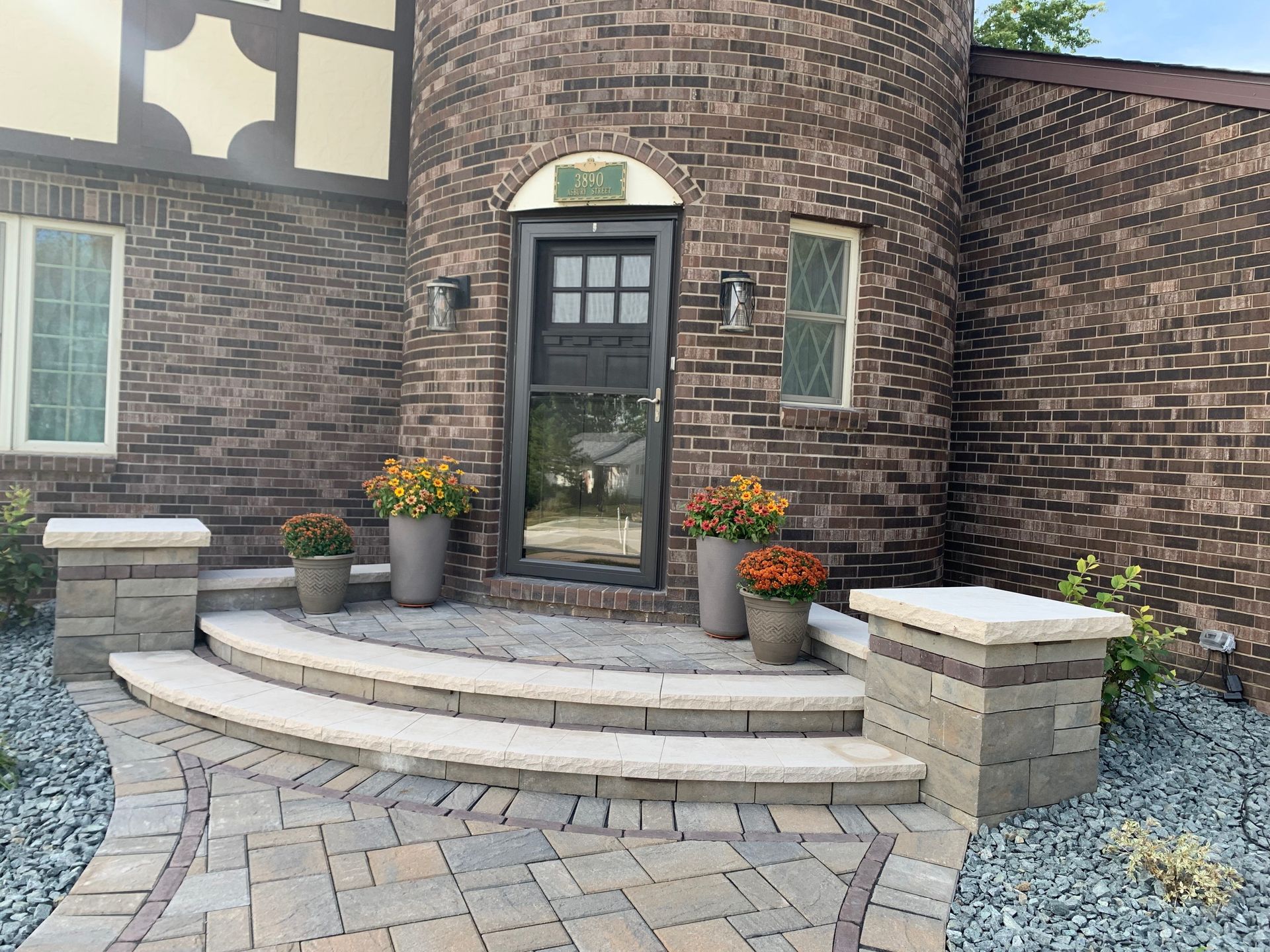 A curved brick home entrance with two steps, stone pillars, potted flowers, and a gray paver walkway.