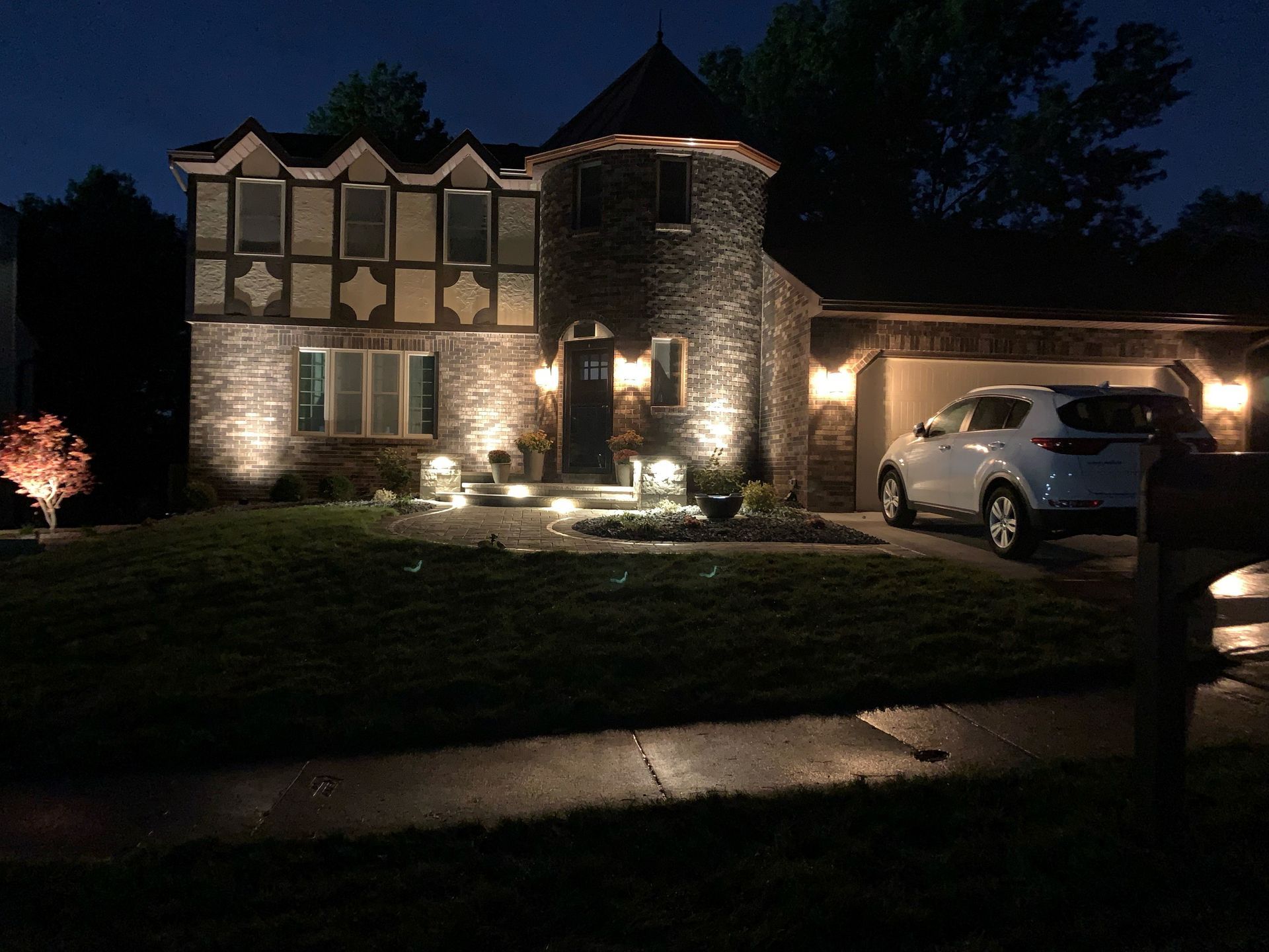 A two-story stone house with a turret at night, illuminated by exterior lights, with a car parked in the driveway.
