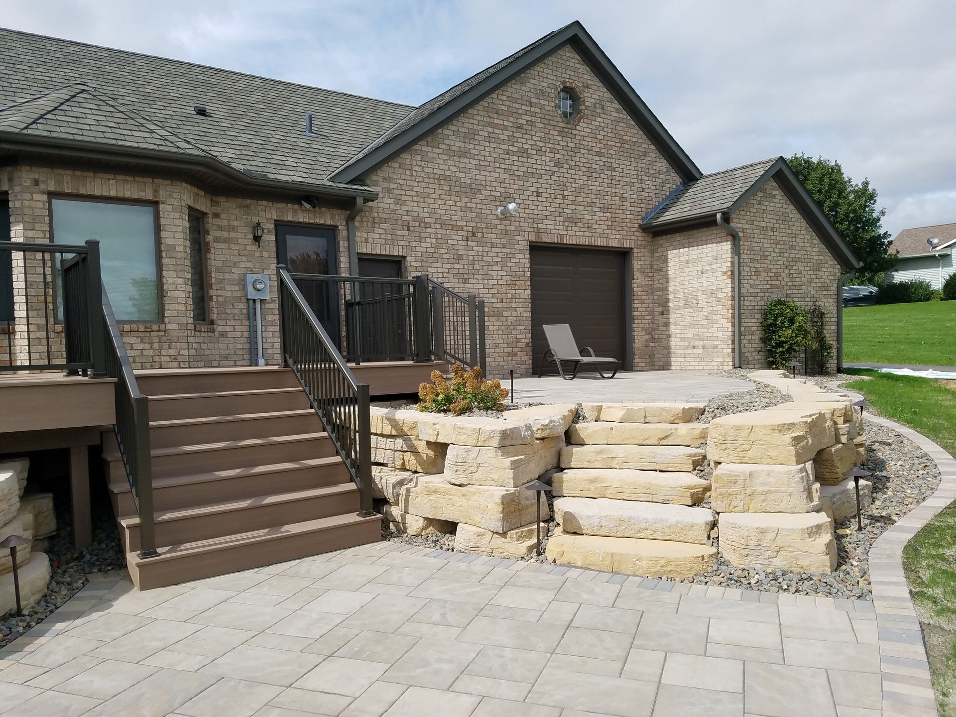 A brick house features a wooden deck with stairs leading down to a stone patio with tiered limestone steps and landscaping.
