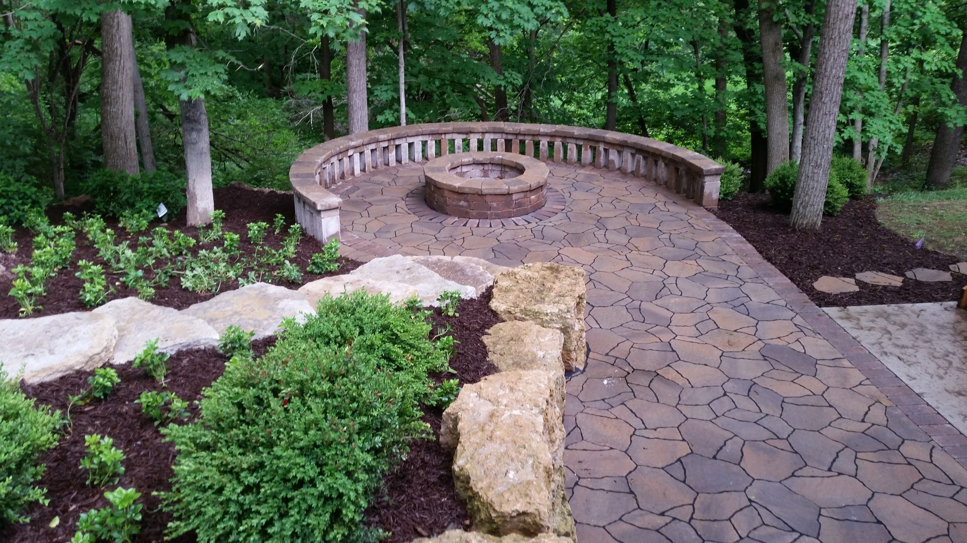 A circular stone fire pit sits on a flagstone patio surrounded by a curved stone bench in a lush, wooded backyard setting.