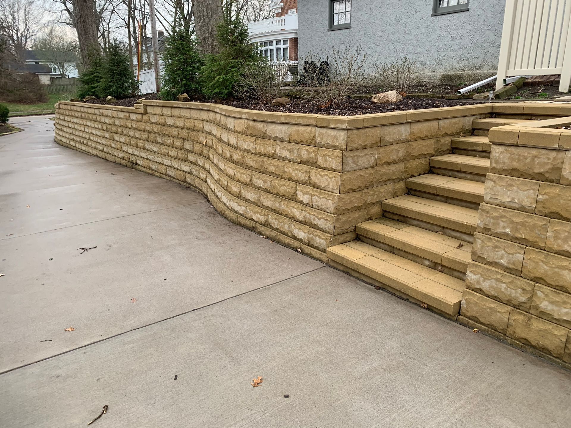 A curved, tan stone retaining wall borders a concrete driveway, featuring built-in stone steps leading to a garden area.