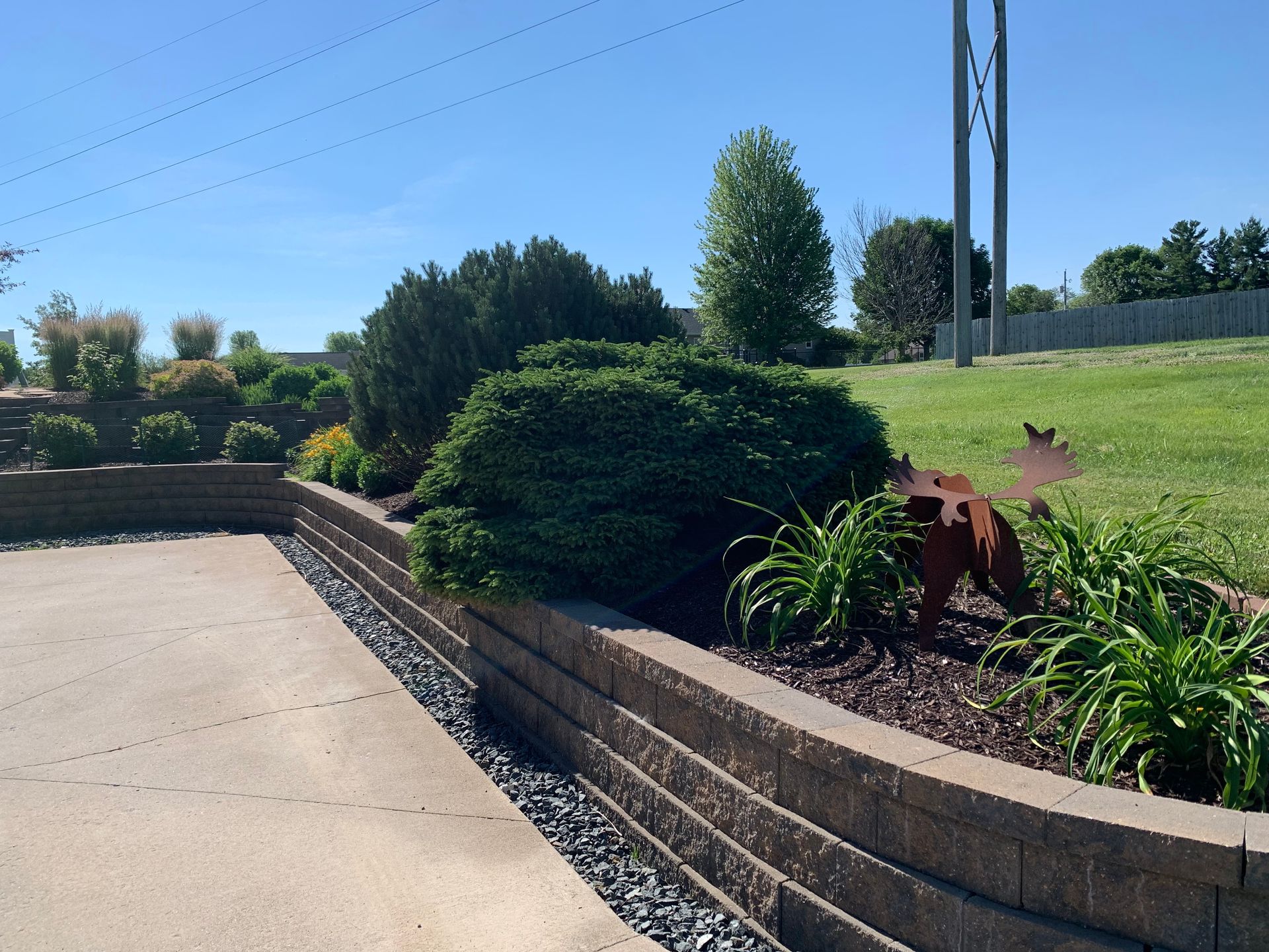 A concrete driveway bordered by a tiered retaining wall, landscaping shrubs, and a metal moose silhouette statue.