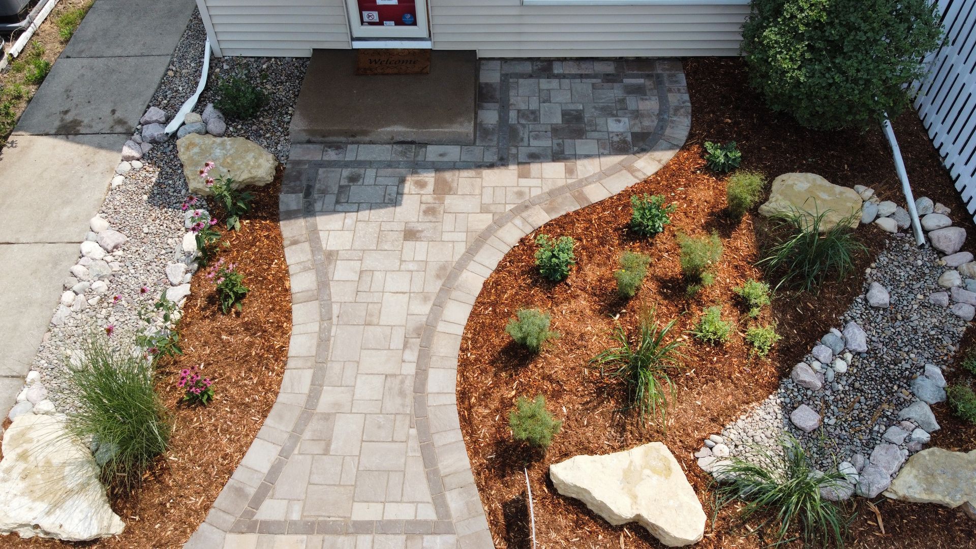 A high-angle view of a front yard featuring a paved stone walkway, mulch garden beds, large decorative rocks, and plants.