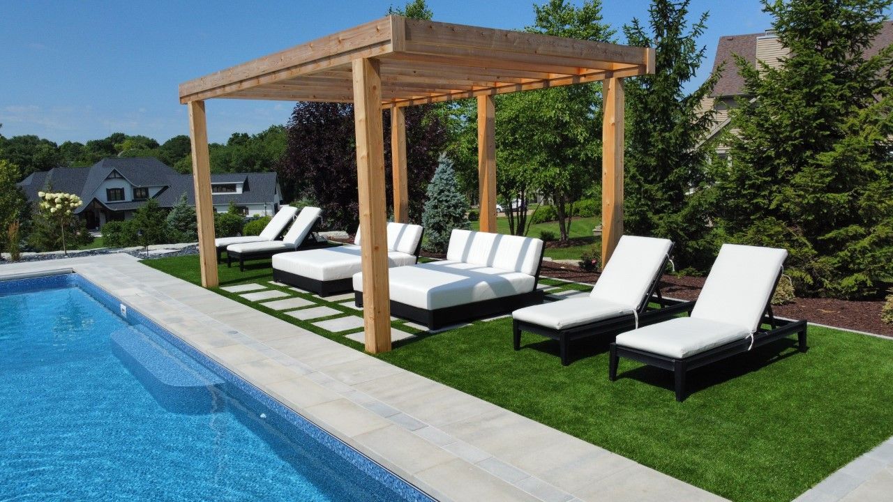 A poolside patio with a wooden pergola, outdoor lounge seating, and several white lounge chairs on a manicured lawn.