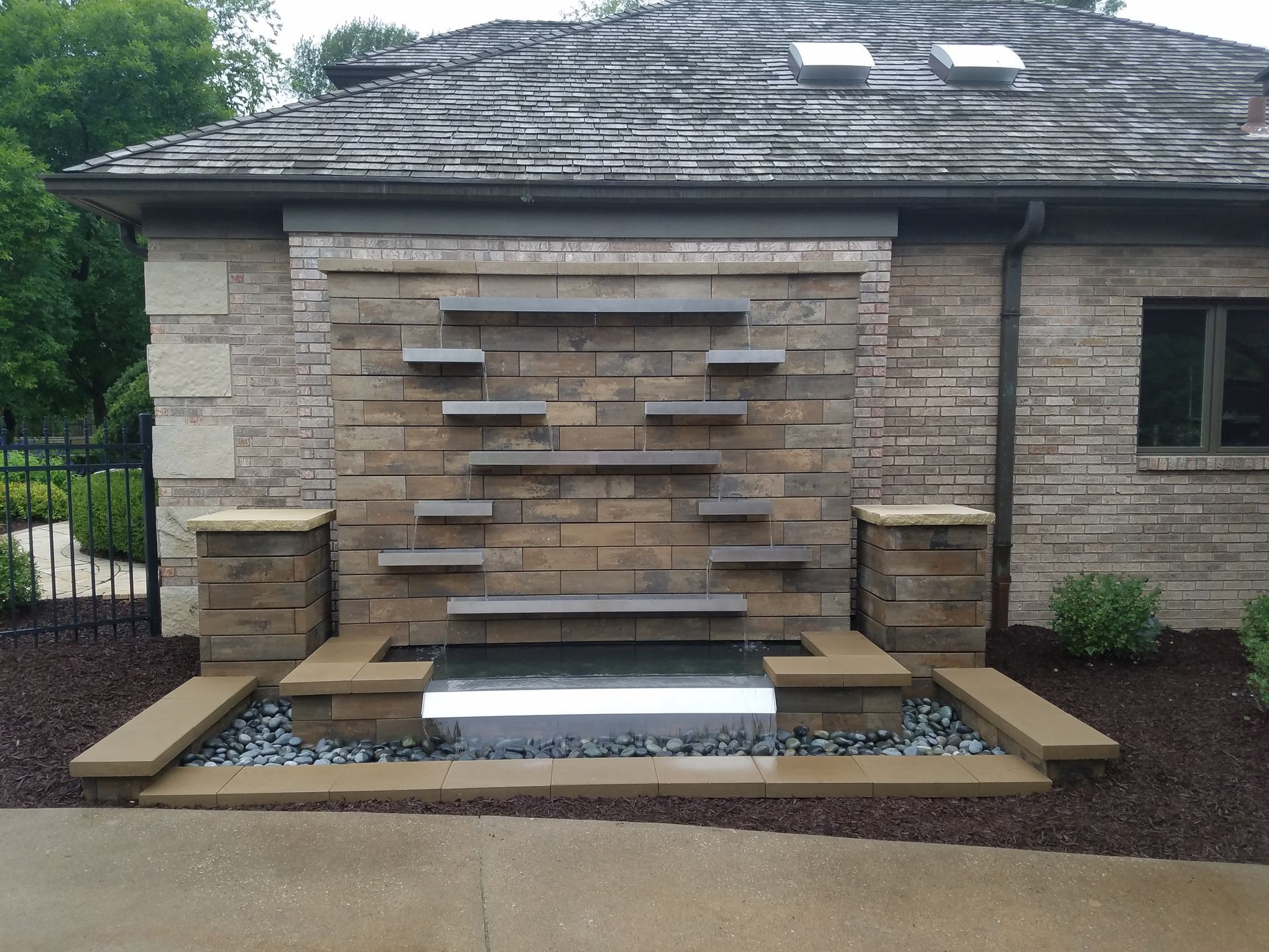 A tiered stone water feature with metal shelves mounted on a brick exterior wall against a house with a shingled roof.