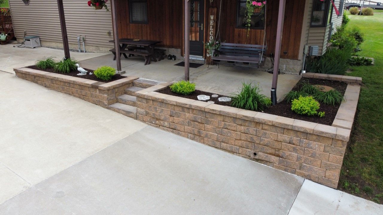 A paved patio area with a tan, stacked-stone retaining wall featuring small green plants and stairs leading to a porch.