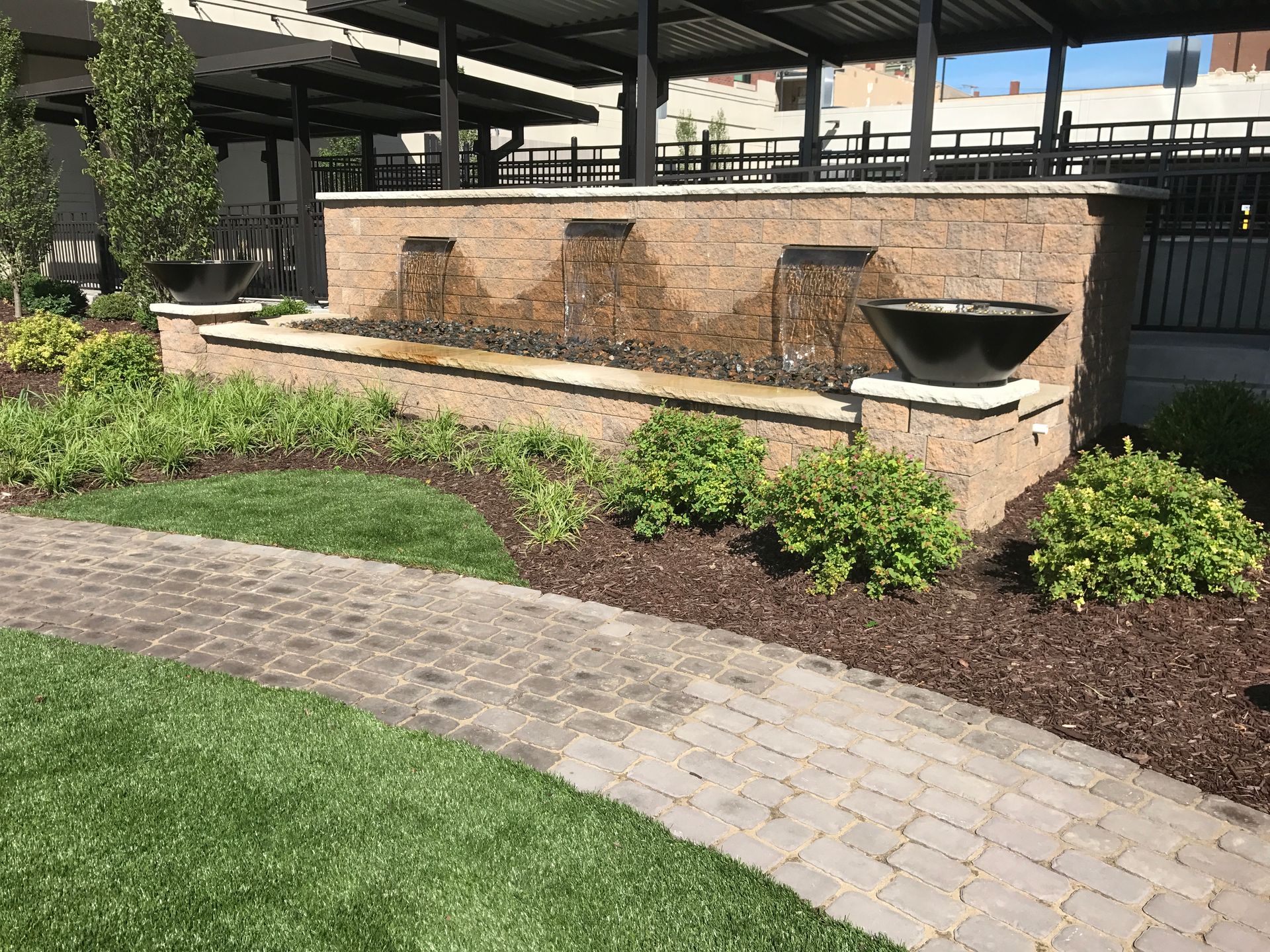 A beige brick water feature with three streams flowing into a basin, set in a garden with pavers and green bushes.