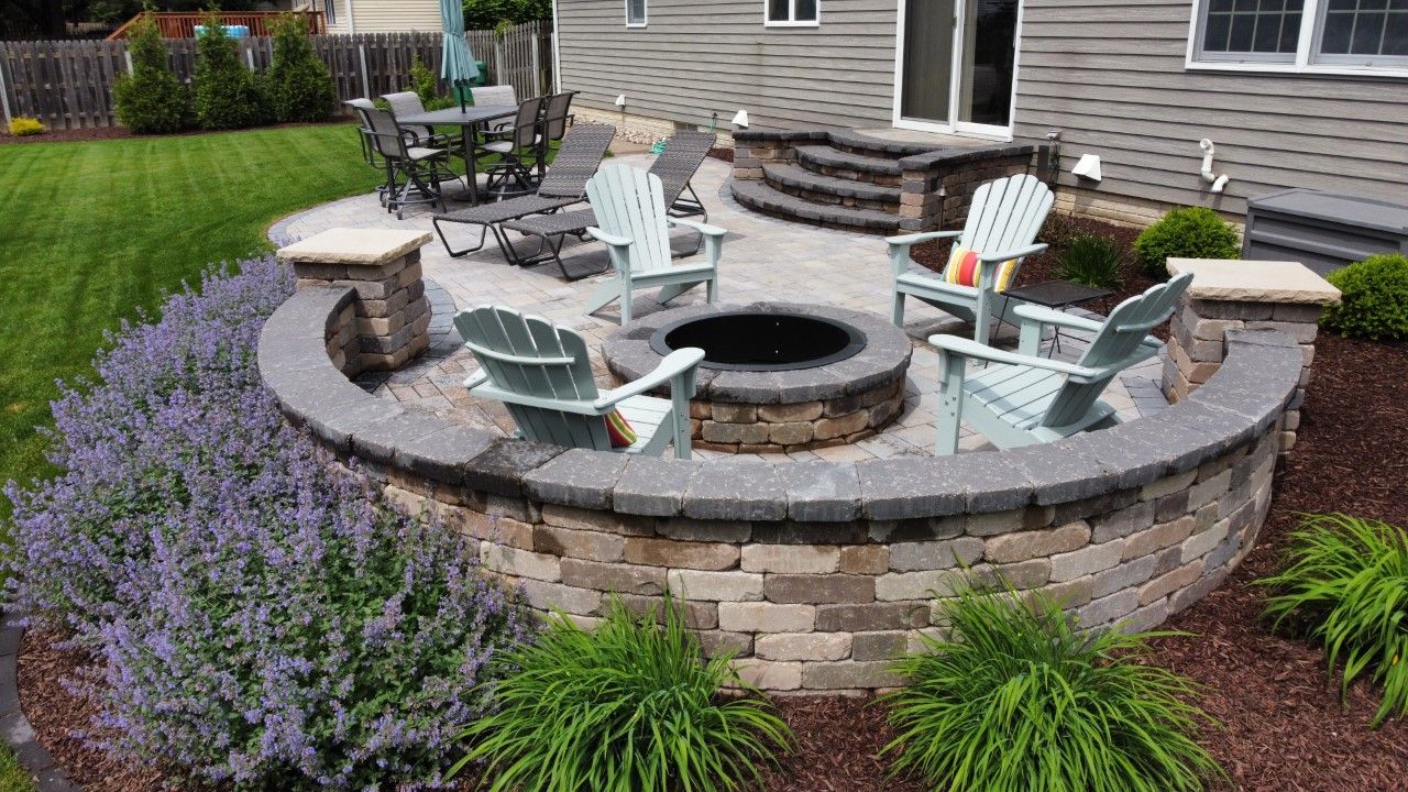 A backyard patio featuring a circular stone fire pit, light blue Adirondack chairs, and a curved retaining wall.