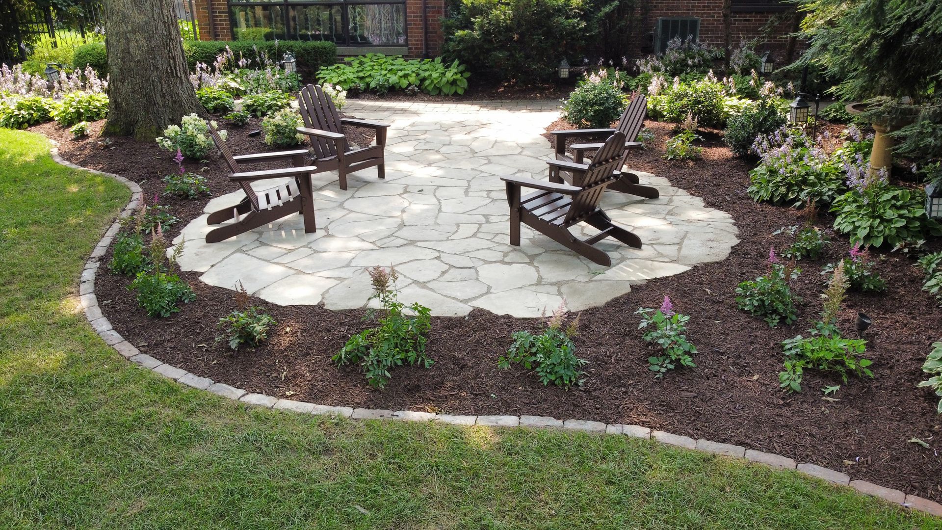 A stone patio with three dark wooden Adirondack chairs, set in a backyard surrounded by mulch and green landscaping.