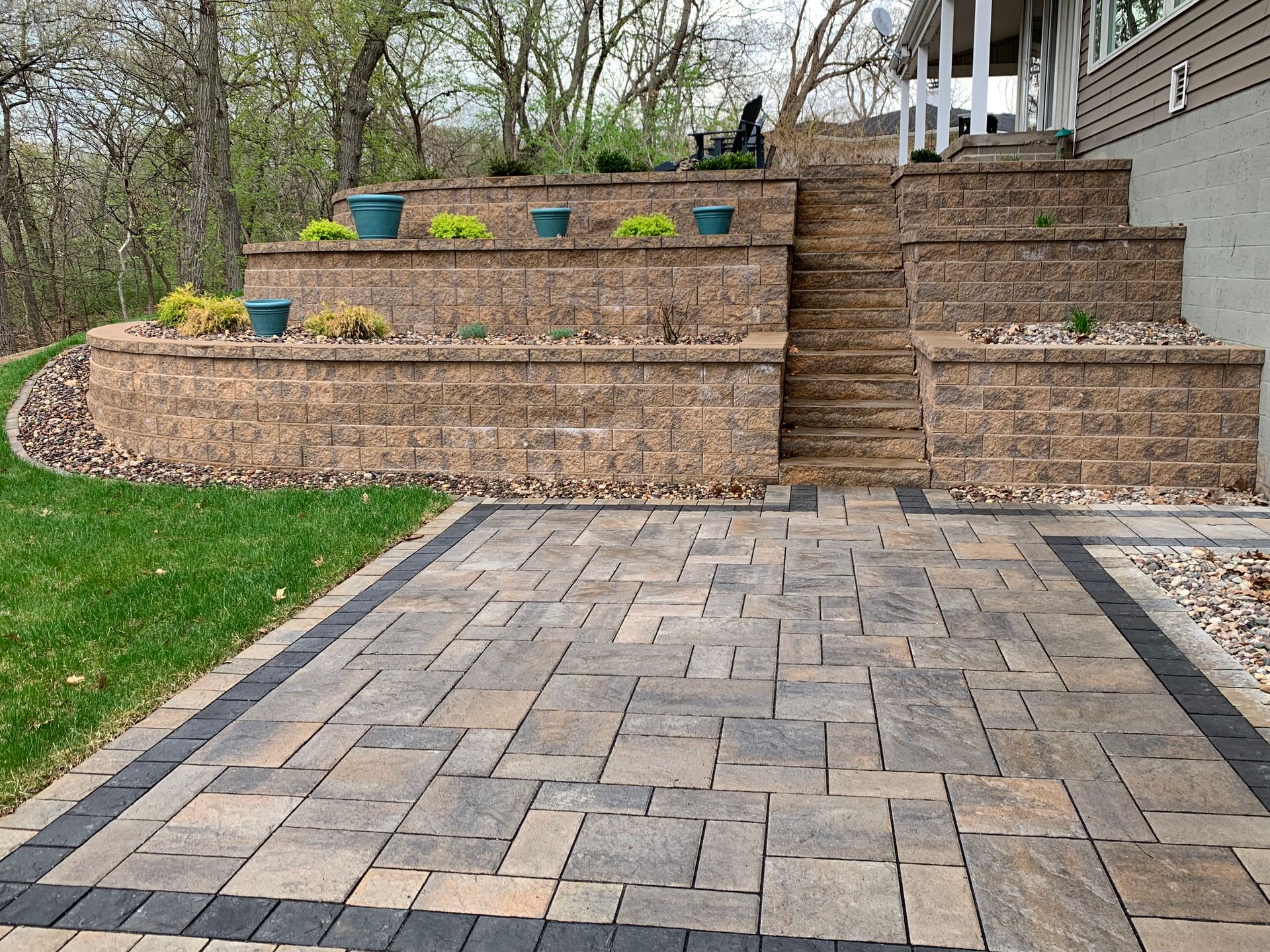 Stone patio with a tan brick retaining wall, tiered garden beds, and stairs leading up to a house exterior.