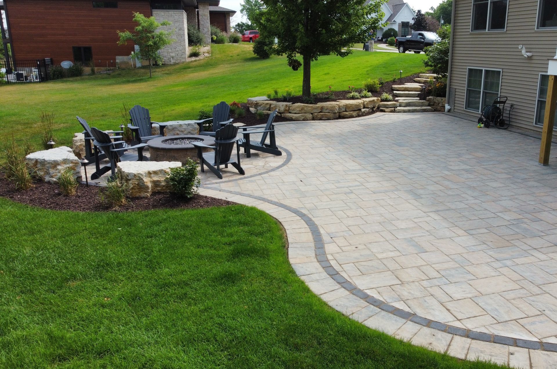 A stone patio with a fire pit surrounded by dark chairs, bordering a lawn and a house on a sunny day.