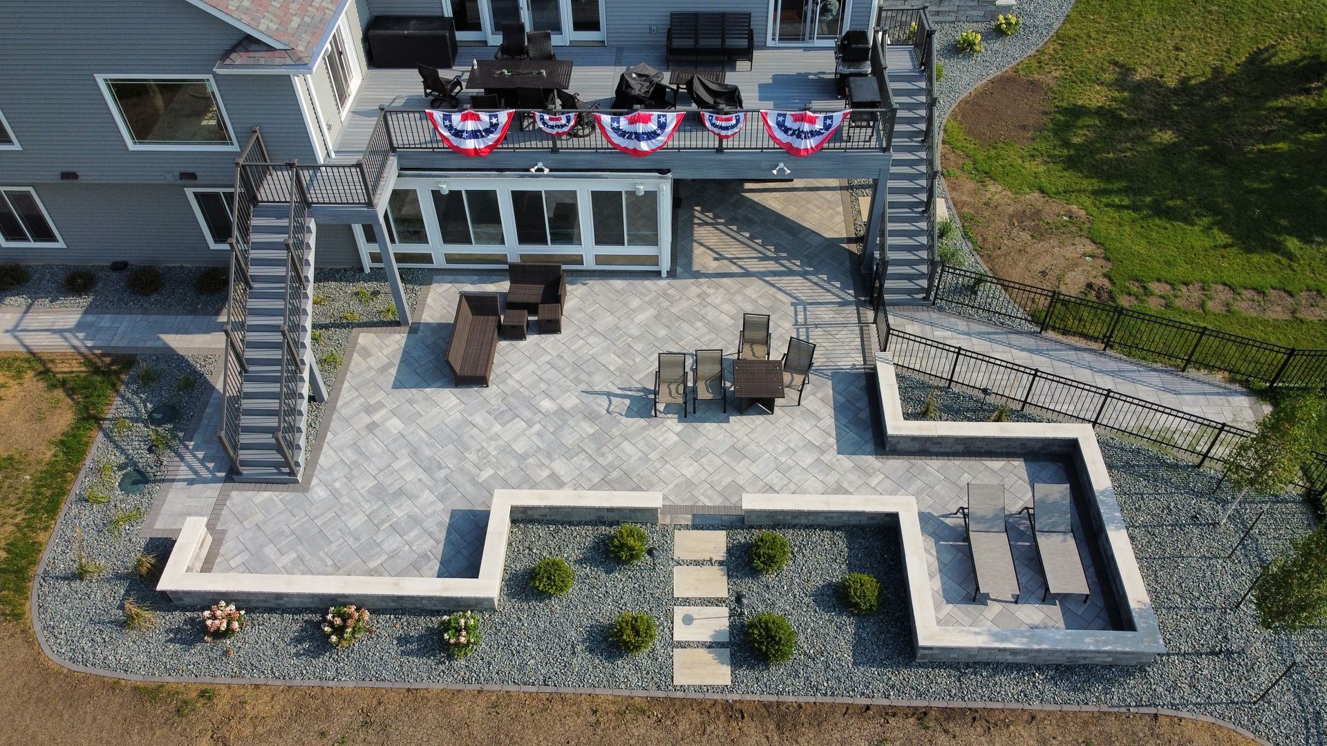 An elevated view of a gray-toned stone patio with dining furniture, lounge chairs, and a retaining wall.