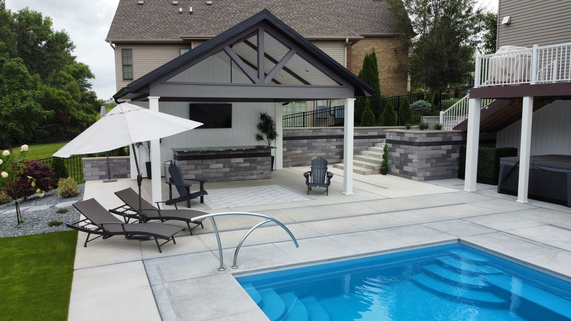 A backyard pool area featuring a covered patio, outdoor kitchen, stone retaining walls, and lounge chairs on a light deck.