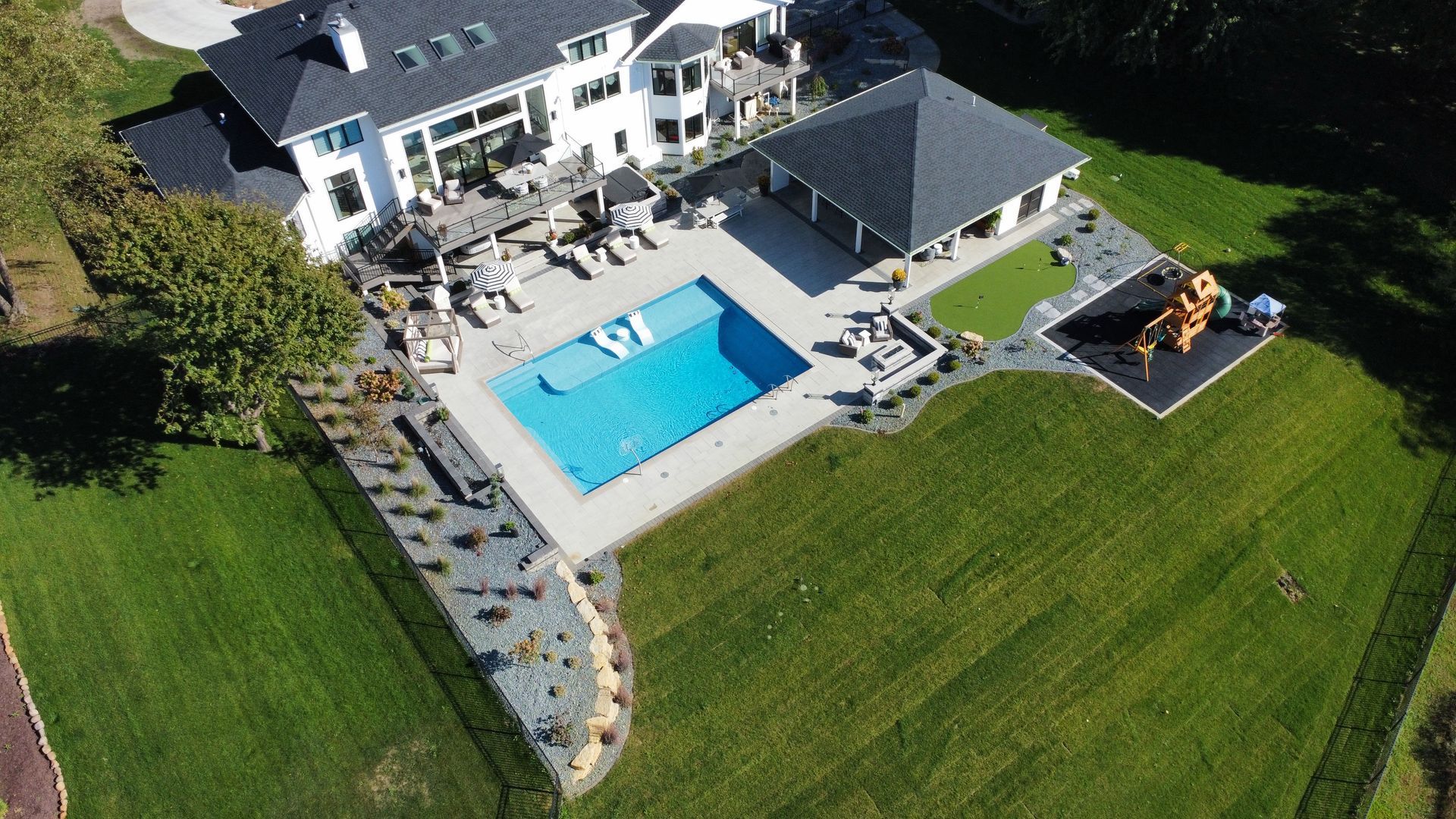 Aerial view of a white house with a backyard pool, patio, cabana, and a children's play set on a lawn.