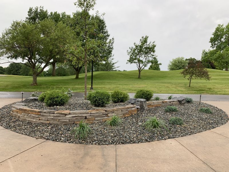A stone garden bed with small shrubs and river rocks sits in a circular concrete plaza against a backdrop of trees.