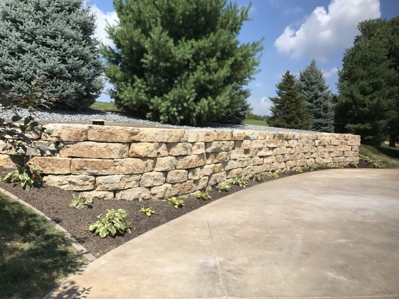 A curved stone retaining wall borders a concrete driveway, set against a backdrop of lush green evergreen trees.