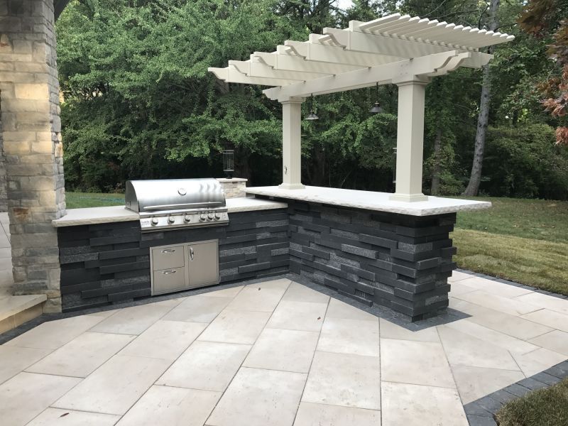 An outdoor kitchen with dark stacked stone, stainless steel grill, light stone countertops, and a white pergola.
