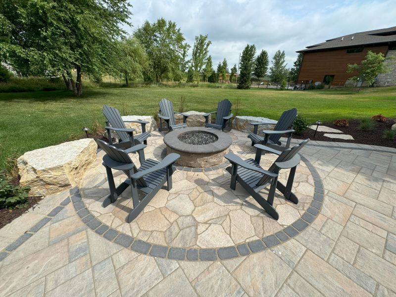 A stone fire pit surrounded by five dark gray Adirondack chairs on a paved patio in a grassy backyard.