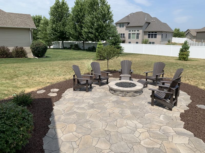A stone patio with a fire pit and Adirondack chairs arranged in a circle in a backyard.