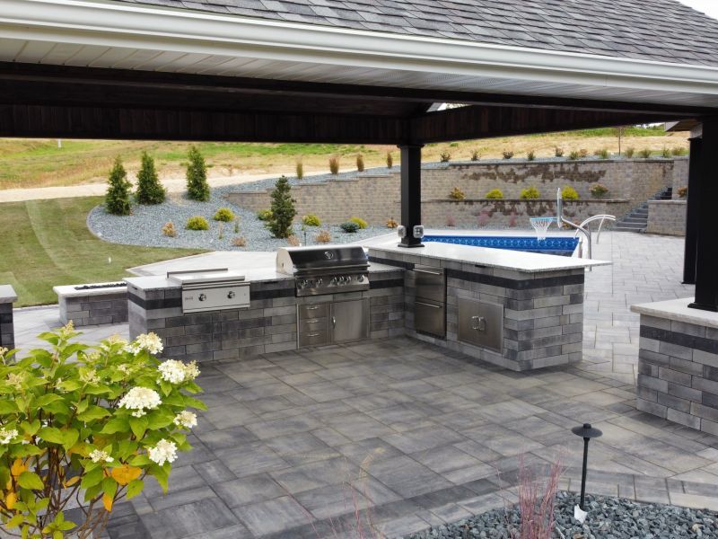 Outdoor kitchen with a grill and countertops under a covered patio, overlooking a pool and landscaped yard.