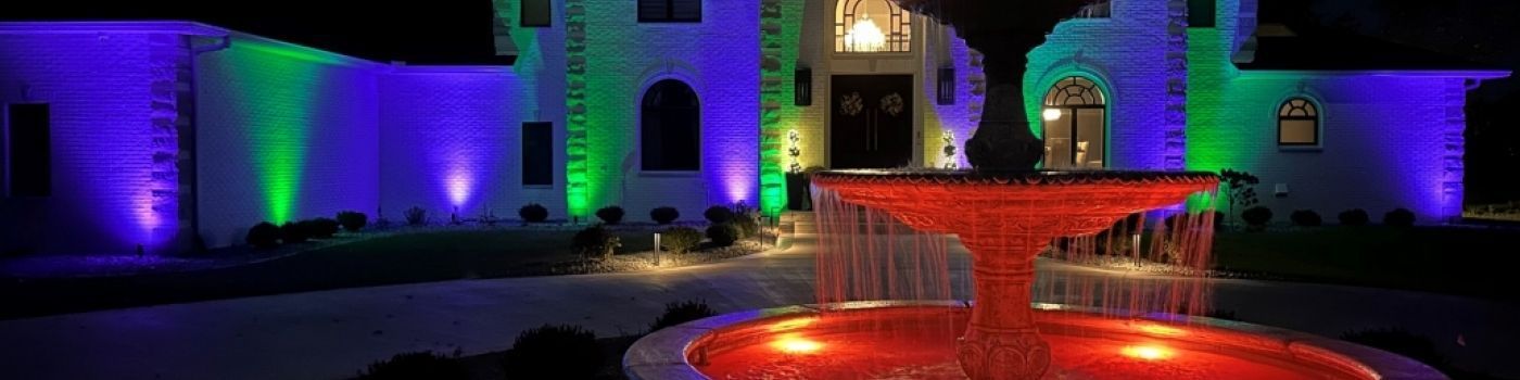 A lit stone fountain glows orange in front of a house illuminated with vibrant green and purple accent lights at night.
