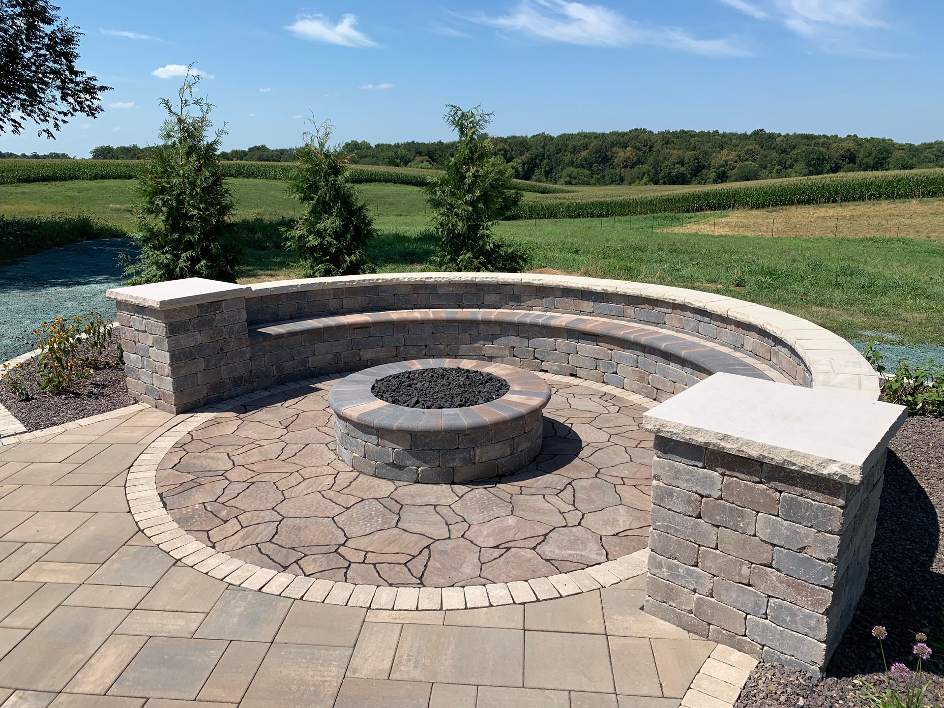 A circular paver patio with a stone fire pit, built-in curved seating, and two stone pillars, set in a rural landscape.