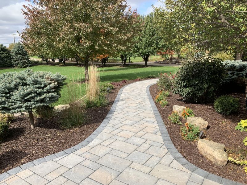 A winding paved walkway in a garden, lined with flowerbeds, leading toward trees in the background on a sunny day.