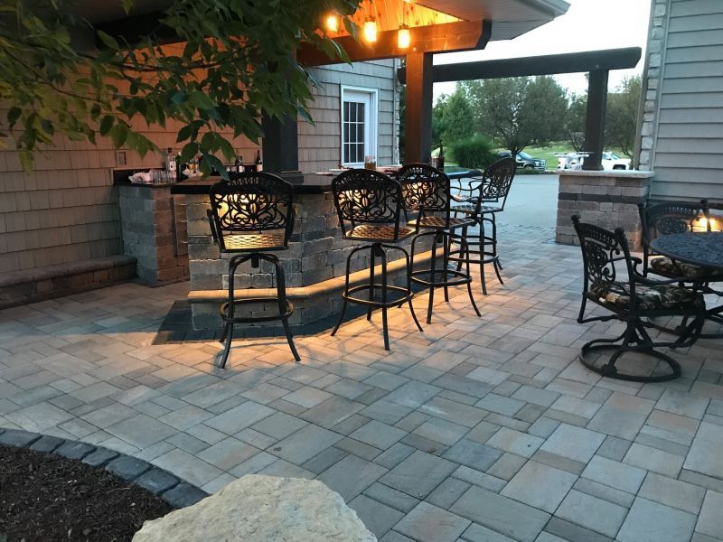 Outdoor bar with bar stools, a stone countertop, and hanging lights. Paved patio with a table and chairs, next to a building.