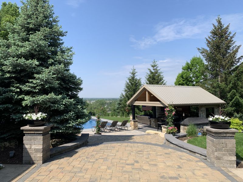 Paver driveway leading to a pool and outdoor kitchen with a view of the surrounding area on a sunny day.
