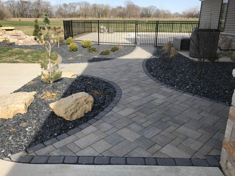 Stone paver pathway curves through a landscaped yard with black rock beds, leading to a black fence and gate.