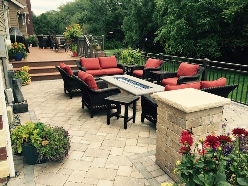 A stone patio with a fire pit, cushioned dark wicker furniture, and potted plants, adjacent to a wooden deck and railing.