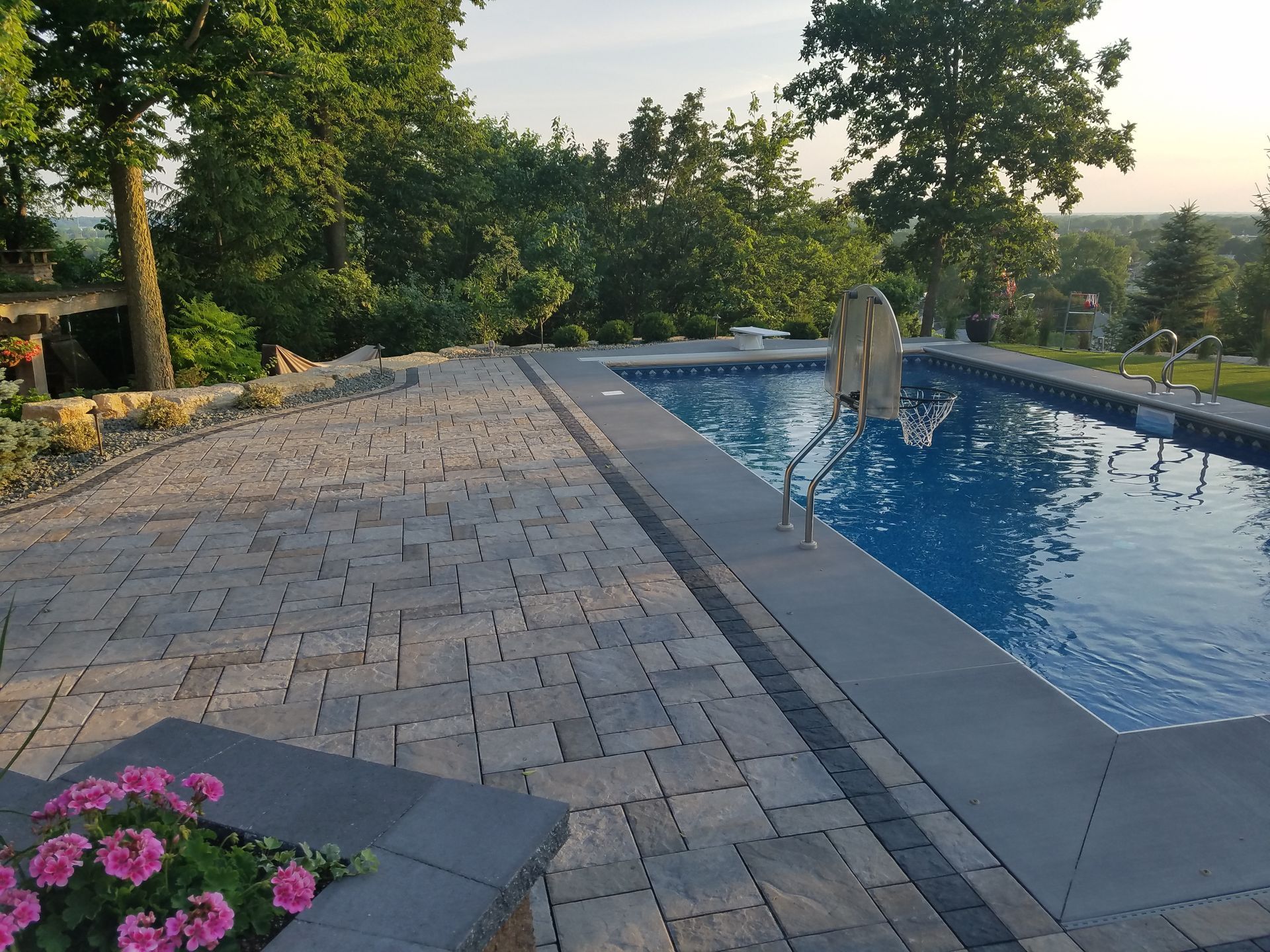 An outdoor patio with stone pavers and a blue swimming pool, overlooking a landscape with trees and distant hills.