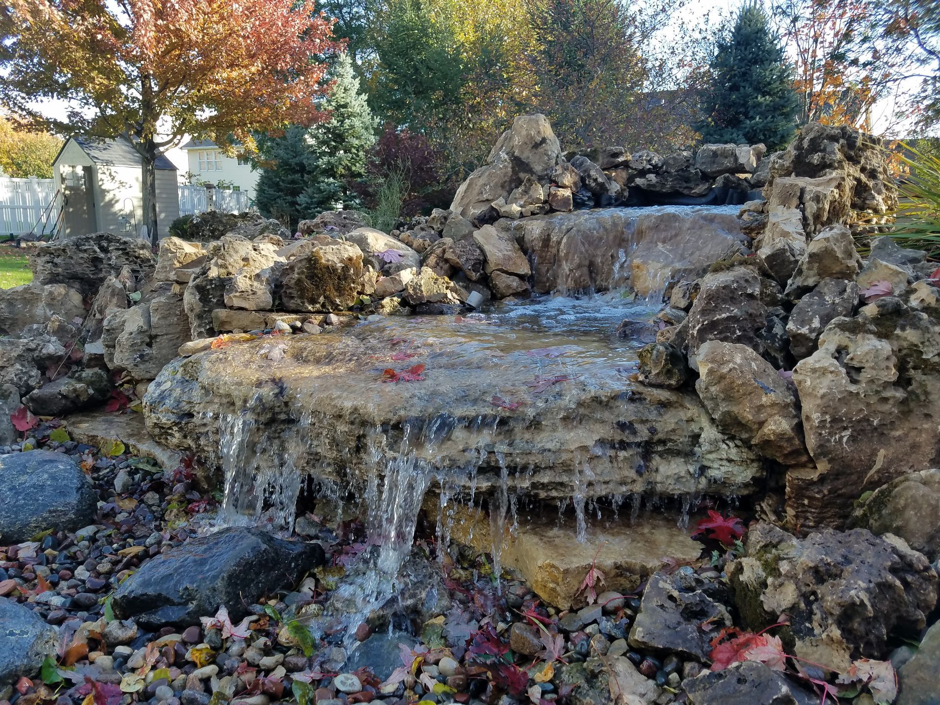 A stone waterfall feature cascading into a rocky pool surrounded by autumn trees and fallen leaves.