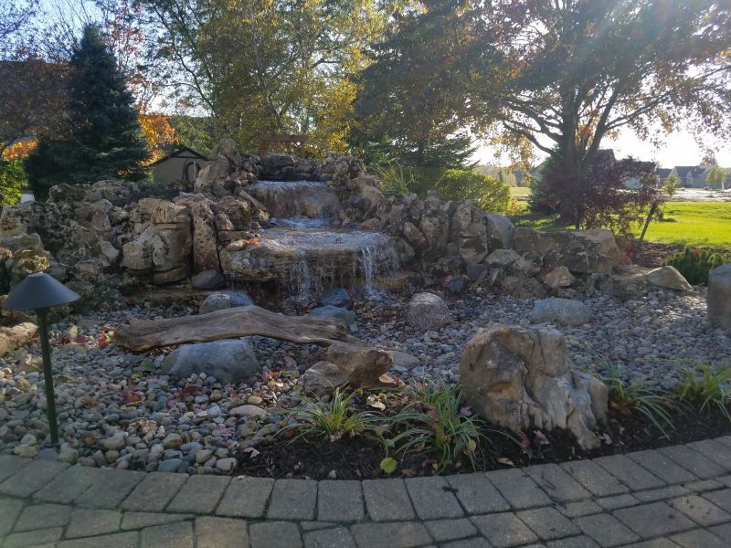 A rock waterfall feature with a small pond, surrounded by landscaping stones and a paved patio area in an outdoor setting.