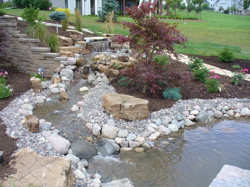 A stone waterfall cascades into a small pond, surrounded by decorative rocks, mulch, and a red Japanese maple tree.