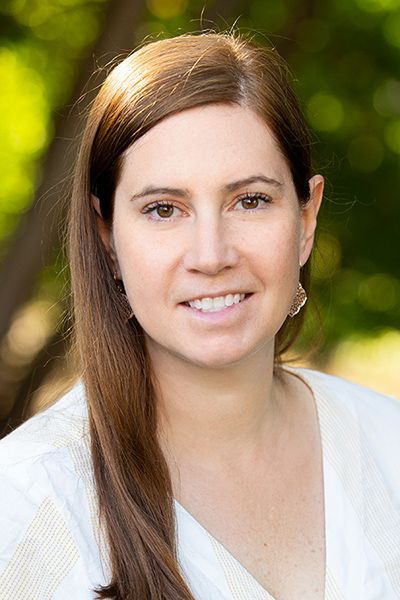 A woman with long brown hair is wearing a white shirt and smiling for the camera.