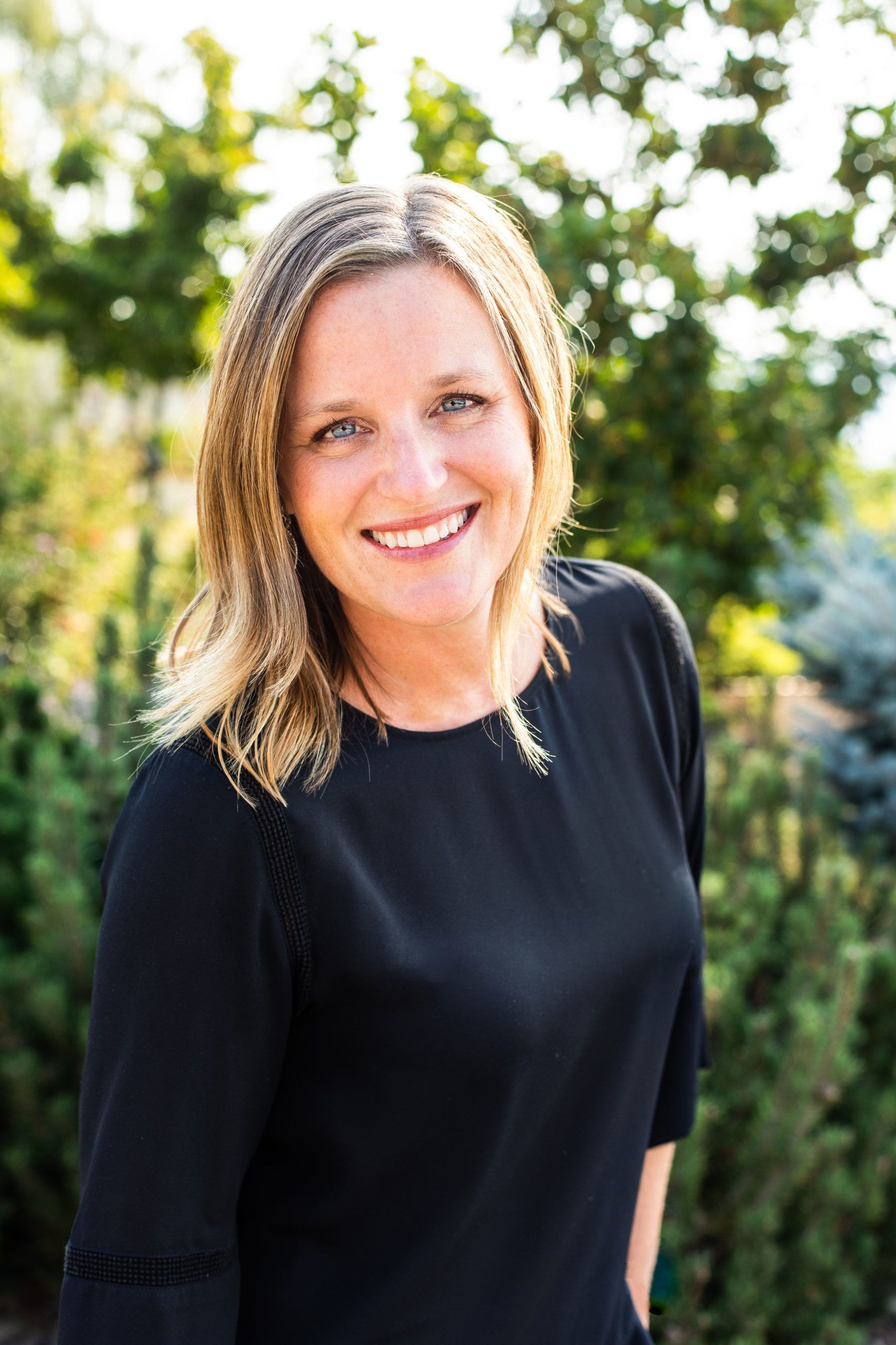 A woman in a black dress is smiling in front of trees.