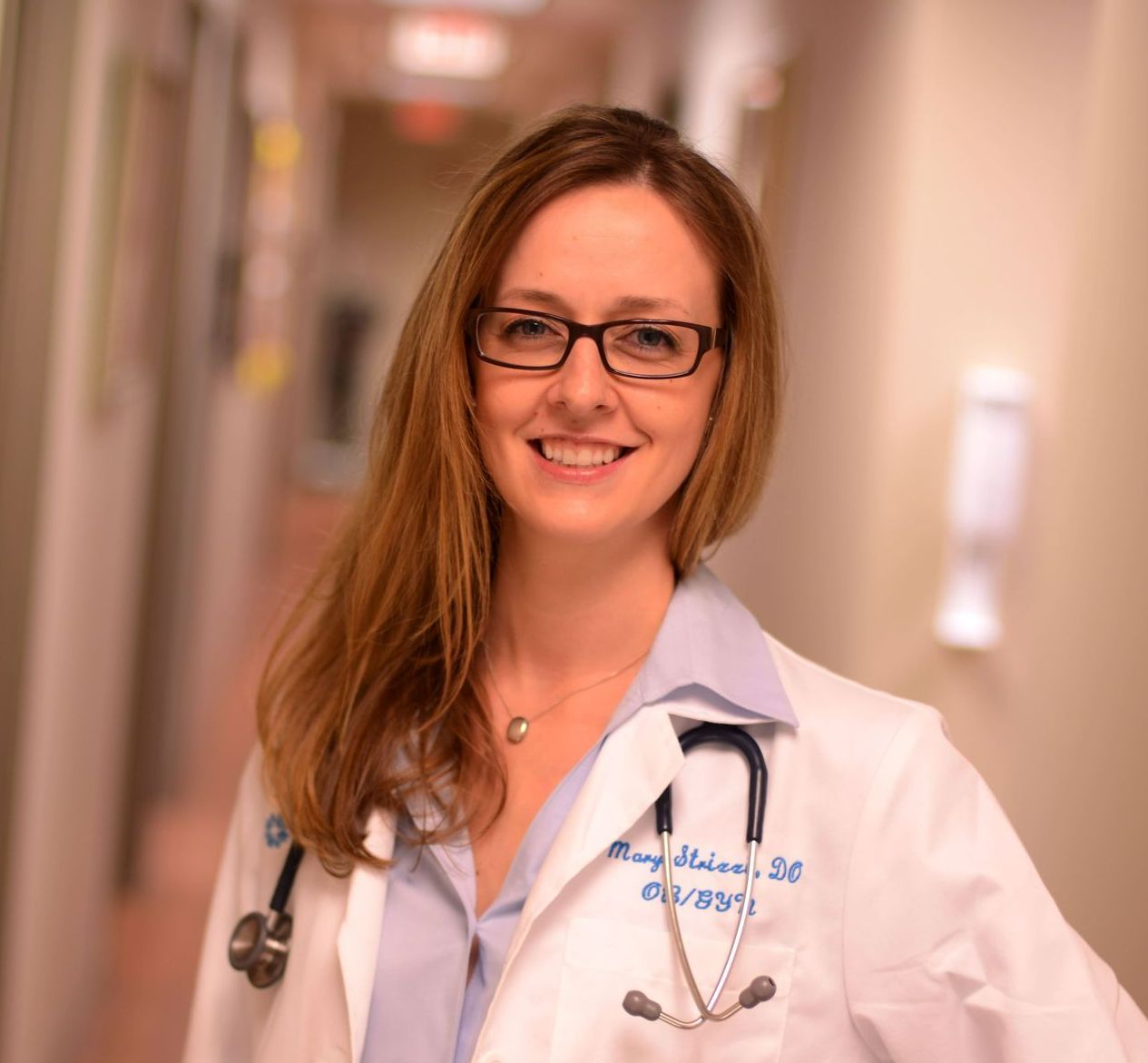 A woman in a lab coat with a stethoscope around her neck is standing in a hallway.