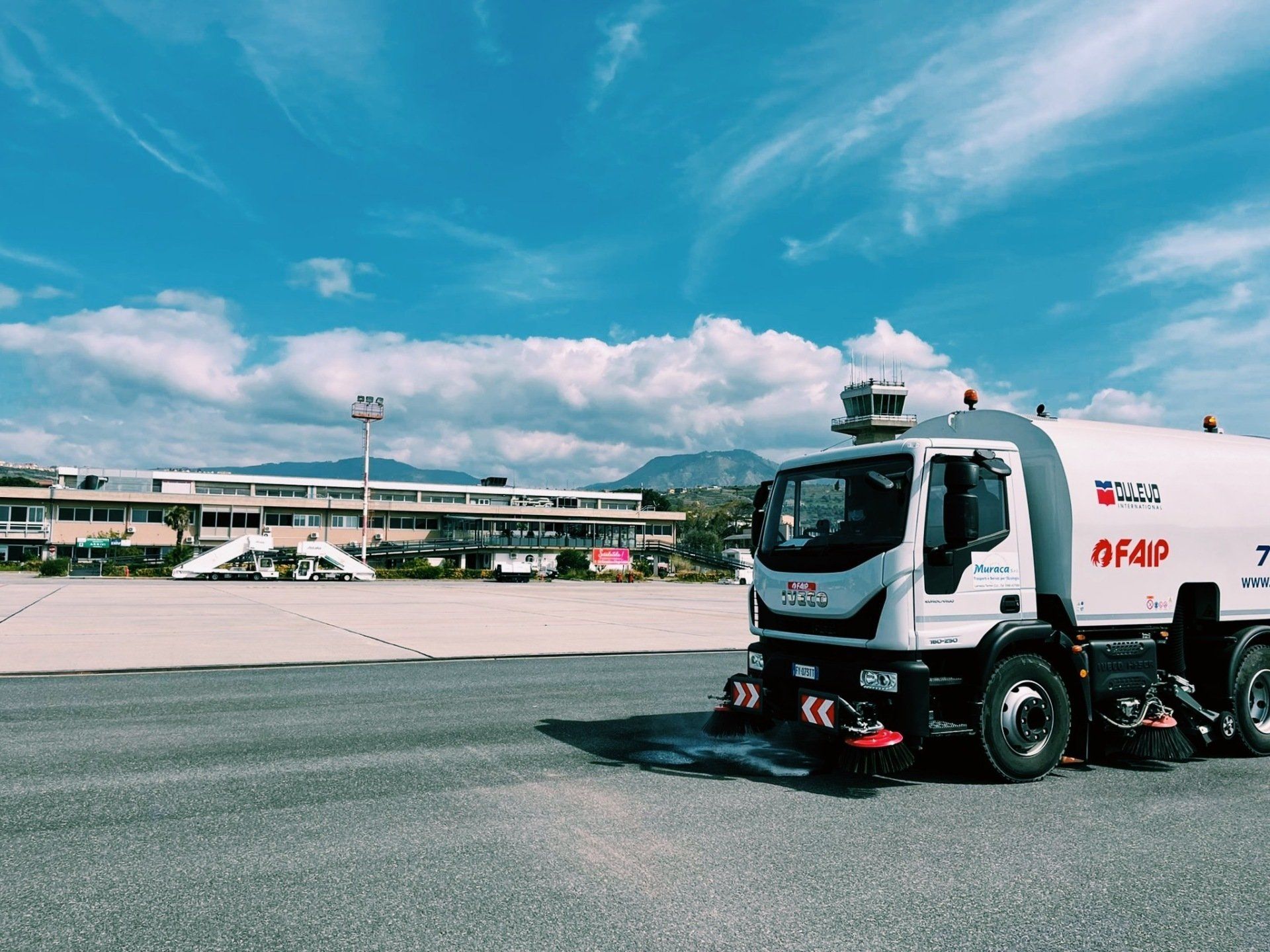 Un camion spazzatrice per piste aeroportuali sull'asfalto sotto un cielo azzurro, con gli edifici del terminal sullo sfondo.